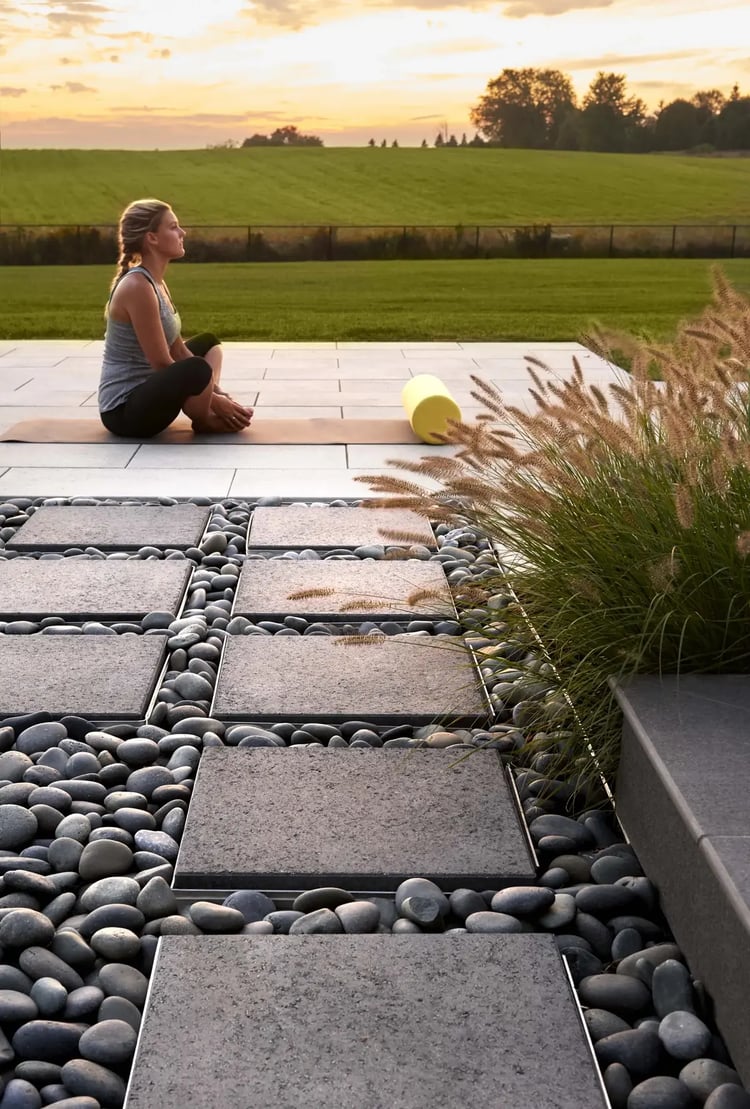 Modern outdoor patio with Techo-Bloc Para slabs and river rock inlays, featuring a yoga session at sunset with open views of a green field and ornamental grasses.