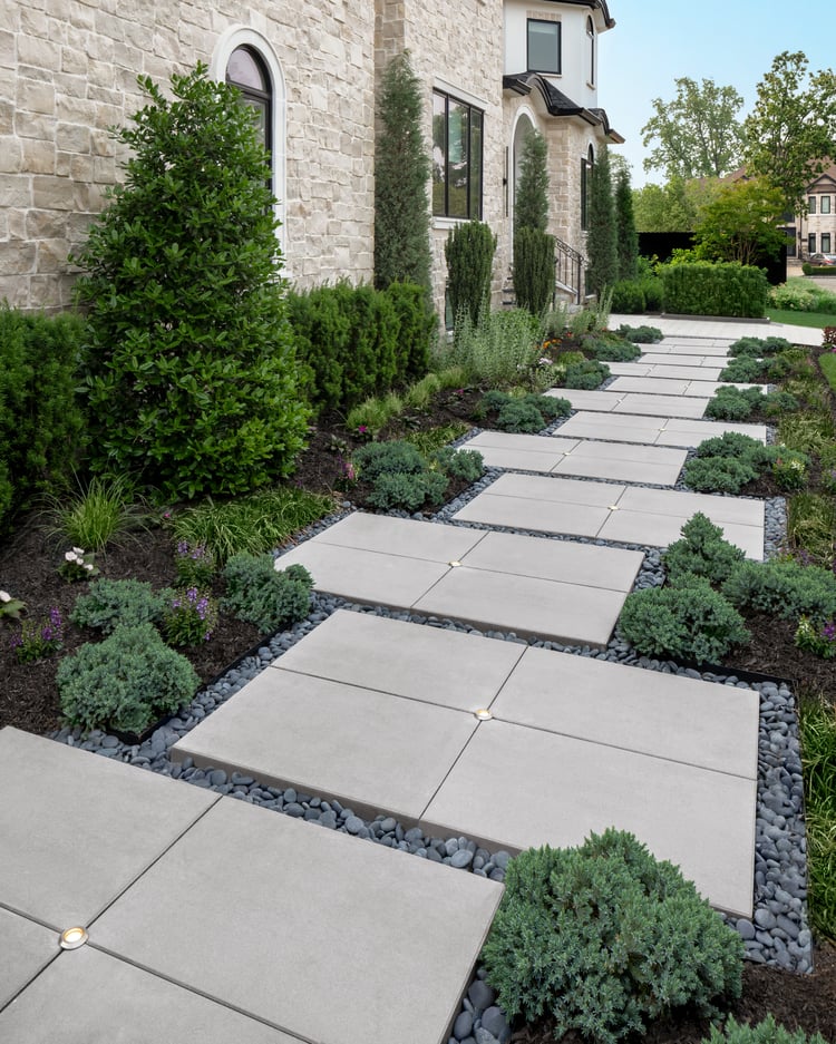 Contemporary front walkway using Techo-Bloc Para slabs with integrated lighting, surrounded by black decorative pebbles and lush evergreen plantings alongside a stone-clad home.