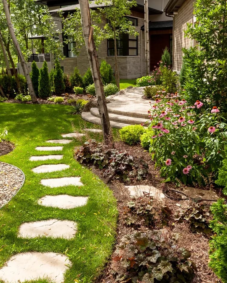 Techo-Bloc Maya stepping stones set in a curved garden path through a lush, flower-filled backyard, leading to a paver landing and front entrance framed by birch trees.